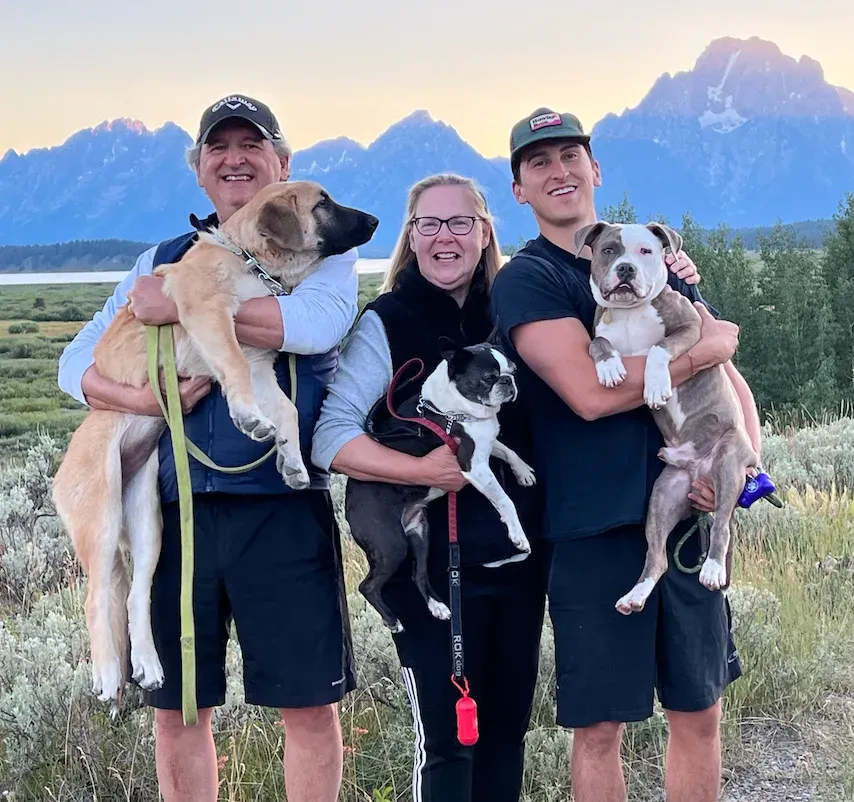 Three team members holding their three dogs together in front of mountains at sunset